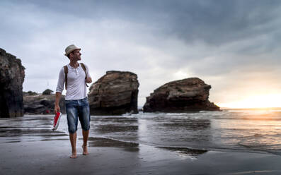Barfüßiger männlicher Rucksacktourist mit Turnschuhen in der Hand, der allein am Sandstrand nahe dem Meer spazieren geht und den Sonnenuntergang bewundert, während er sich nach einer Wanderung in der Natur entspannt - ADSF20033