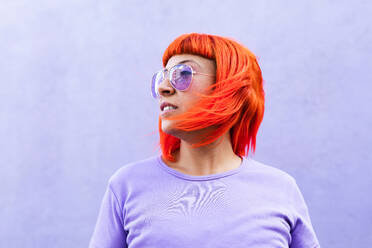 Adult female with vibrant red dyed hair and in stylish sunglasses looking away against violet wall on street - ADSF20034