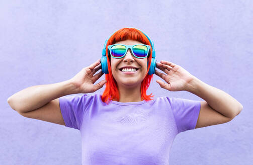 Happy stylish adult female with ginger dyed hair listening to music in headphones and singing along against violet wall in city - ADSF20036