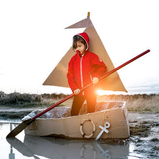 Cute kid with paddle playing in handmade cardboard boat while entertaining in countryside at weekend - ADSF20041