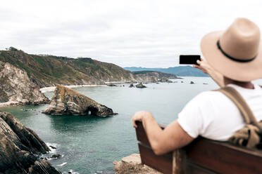 Rückansicht eines männlichen Touristen, der auf einer Bank sitzt und ein Foto von der beeindruckenden Klippe und dem Meer macht, während er seine Reise genießt und sein Smartphone benutzt - ADSF20044
