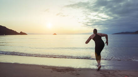 Rückenansicht eines nicht erkennbaren Profischwimmers in Badeanzug und Kappe, bereit zum Laufen im Meerwasser während des Trainings am Meer bei Sonnenuntergang - ADSF20047