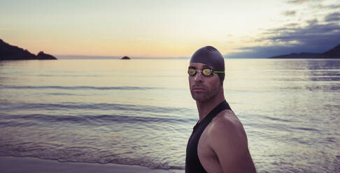 Seitenansicht eines selbstbewussten männlichen Schwimmers mit Kappe und Schwimmbrille, der am Strand vor dem Hintergrund des Meeres unter dem Sonnenuntergang steht und in die Kamera schaut - ADSF20048