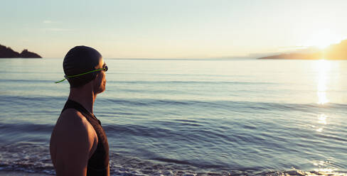 Seitenansicht eines selbstbewussten männlichen Schwimmers mit Kappe und Schwimmbrille, der am Strand vor dem Hintergrund des Meeres bei Sonnenuntergang steht und wegschaut - ADSF20049