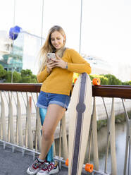 Delighted female skater with bionic leg prosthesis standing on bridge with longboard and reading messages on social media on smartphone - ADSF20070