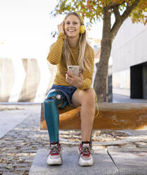 Front view of smiling female with bionic leg prosthesis sitting in street and enjoying music in headphones on sunny day - ADSF20072