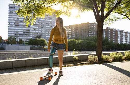 Optimistische Skaterin mit Beinprothese, die auf einem Longboard in einem Stadtpark steht und wegschaut, während sie den sonnigen Tag genießt - ADSF20073