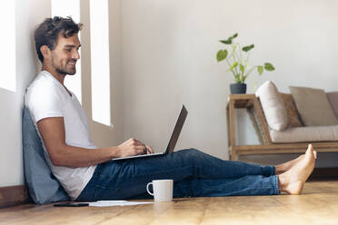 Smiling man using laptop while sitting on floor at home - SBOF02362