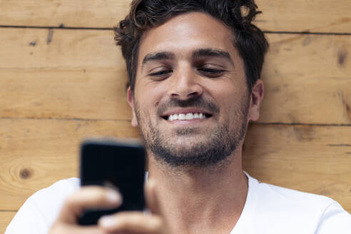 Lächelnder Mann, der ein Mobiltelefon benutzt, während er zu Hause auf dem Boden liegt - SBOF02365