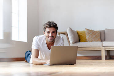 Mittlerer erwachsener Mann lächelt, während er einen Laptop benutzt, der zu Hause auf dem Boden liegt - SBOF02371