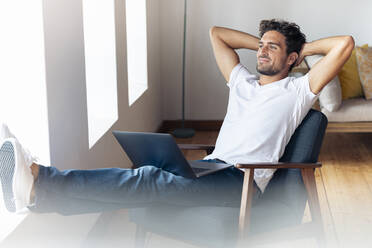 Man with hands behind head relaxing while sitting on armchair at home - SBOF02372