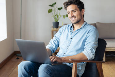 Smiling man using laptop while sitting on armchair at home - SBOF02376