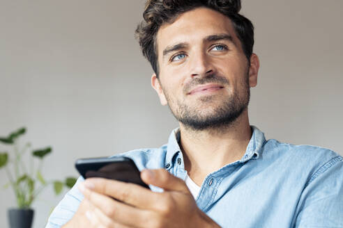 Mann mit blauen Augen, der wegschaut, während er zu Hause ein Mobiltelefon benutzt - SBOF02377