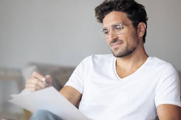 Smiling man wearing eyeglasses writing in paper while sitting at home - SBOF02383