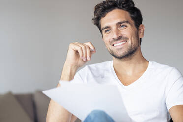 Mid adult man with paper smiling while looking away sitting at home - SBOF02385