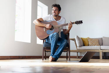 Smiling man playing guitar while sitting on chair at home - SBOF02390