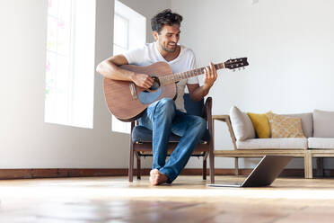 Mittlerer erwachsener Mann übt Gitarre, während er zu Hause auf einem Stuhl neben einem Laptop sitzt - SBOF02391