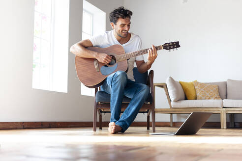 Mid adult man practicing guitar while sitting on chair by laptop at home - SBOF02391