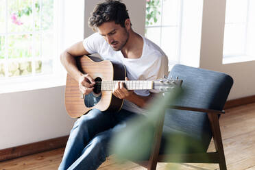 Man practicing on guitar while sitting on armchair at home - SBOF02392