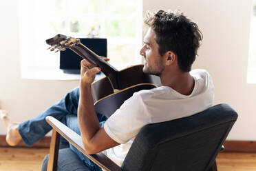 Smiling man playing guitar while relaxing on armchair at home - SBOF02394
