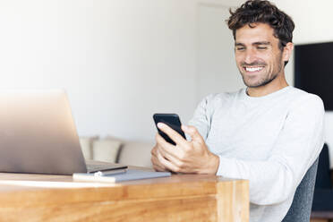 Smiling man with laptop using mobile phone while sitting by table at home - SBOF02406