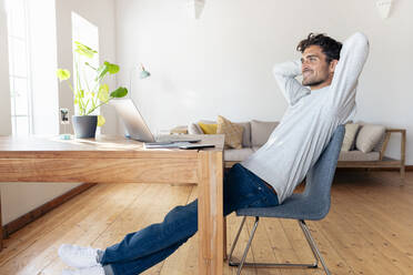 Smiling man with hands behind head relaxing while sitting on chair at home - SBOF02408
