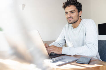 Mid adult man working on laptop while sitting by table at home - SBOF02409