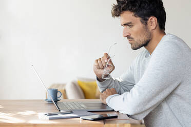 Mann hält eine Brille, während er zu Hause am Laptop arbeitet - SBOF02410