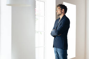 Smiling businessman looking through window while standing with arms crossed at home - SBOF02421