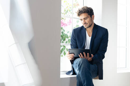 Geschäftsmann, der ein digitales Tablet benutzt, während er zu Hause auf der Fensterbank sitzt - SBOF02422