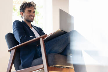 Smiling businessman using laptop while sitting on armchair at home - SBOF02424