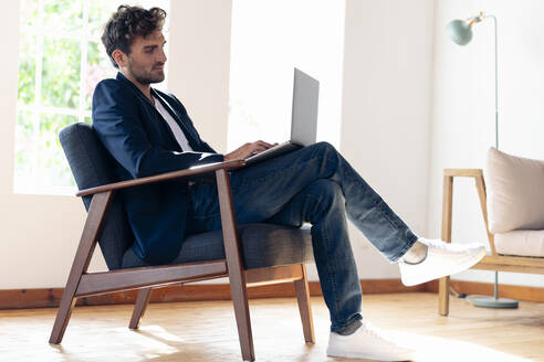 Geschäftsmann arbeitet am Laptop, während er zu Hause auf einem Sessel sitzt - SBOF02426
