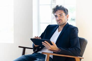 Thoughtful businessman with digital tablet looking away while sitting on armchair at home - SBOF02433
