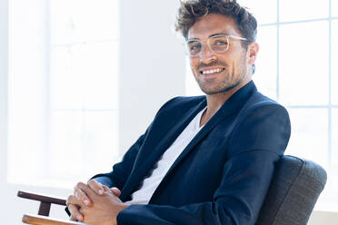 Selbstbewusster Geschäftsmann mit Brille sitzt mit gefalteten Händen auf einem Sessel zu Hause - SBOF02434