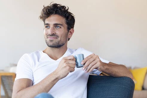 Lächelnder Mann trinkt Kaffee und sitzt auf einem Stuhl zu Hause - SBOF02436
