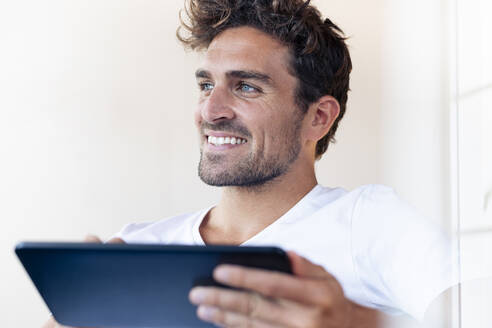 Lächelnder Mann mit digitalem Tablet, der wegschaut, während er zu Hause sitzt - SBOF02437