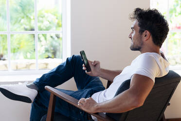 Mann mit Smartphone schaut durch das Fenster, während er sich zu Hause auf einem Sessel entspannt - SBOF02444