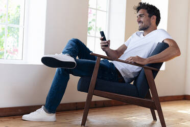 Smiling man using smart phone while resting on armchair at home - SBOF02445