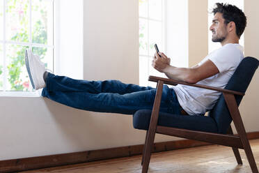 Man with smart phone looking through window while relaxing on armchair at home - SBOF02447