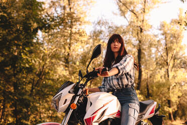 Confident young woman riding motorcycle amidst trees in forest - CAVF91690
