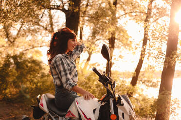 Happy young woman relaxing sitting on motorcycle amidst trees - CAVF91691