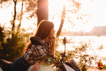 Junge Frau, die auf einem Motorrad sitzend die Aussicht betrachtet und sich am Fluss entspannt - CAVF91692