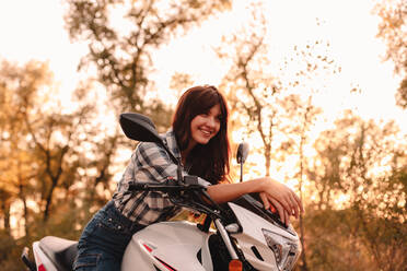 Porträt einer glücklichen jungen Frau, die auf einem Motorrad inmitten von Bäumen sitzt - CAVF91695