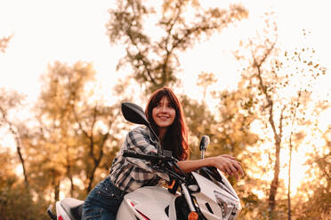Porträt einer glücklichen jungen Frau, die auf einem Motorrad inmitten von Bäumen sitzt - CAVF91696