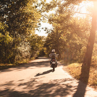 Rückansicht einer Motorrad fahrenden Frau auf einer Landstraße im Wald - CAVF91697
