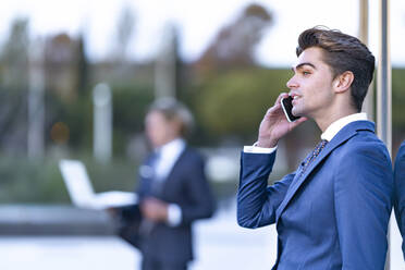 Junger Geschäftsmann, der mit seinem Handy telefoniert und sich an ein gläsernes Bürogebäude lehnt - GGGF00864