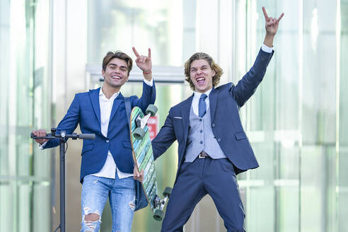 Cheerful young male coworkers making shaka sign against office building - GGGF00870