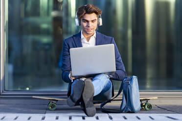 Junger Geschäftsmann arbeitet am Laptop und hört Musik auf dem Skateboard sitzend an einem sonnigen Tag - GGGF00874