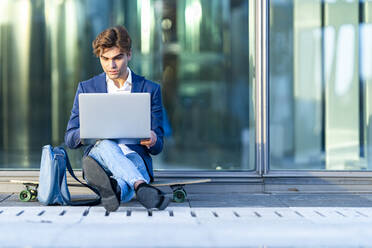 Männlicher Unternehmer, der einen Laptop benutzt, während er auf einem Skateboard sitzt und an einem sonnigen Tag gegen ein Glas lehnt - GGGF00875