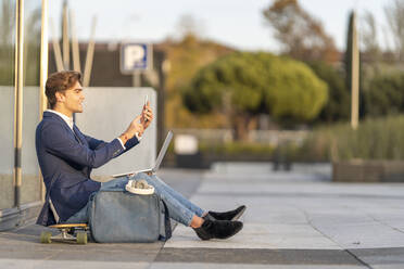 Fröhlicher Geschäftsmann mit Laptop bei einem Videogespräch auf dem Skateboard sitzend an einem sonnigen Tag - GGGF00877